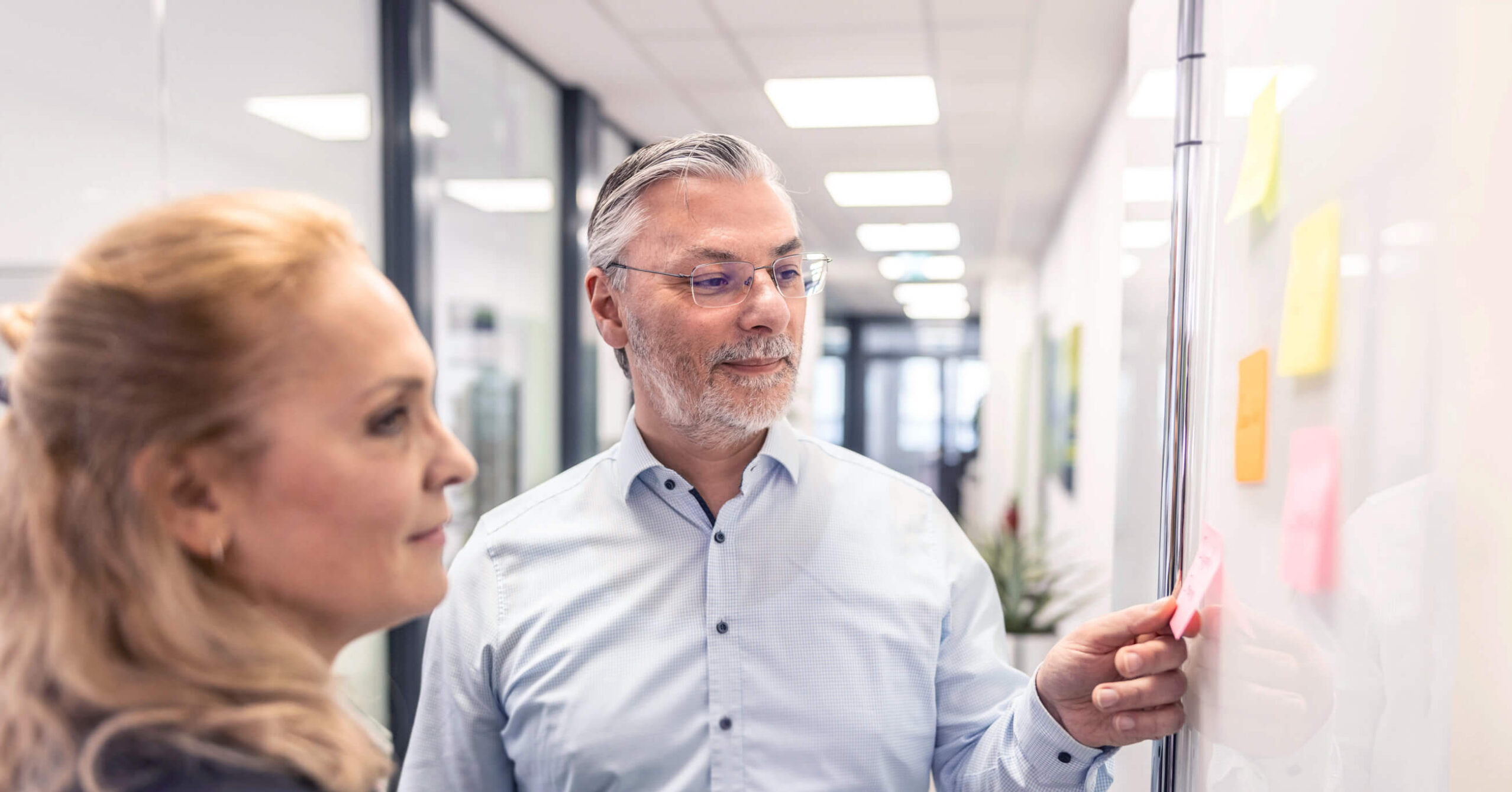 Eine HSP-Mitarbeitende und ein Kunde stehen vor einem Whiteboard mit farbigen Post-its und besprechen gemeinsam Inhalte.